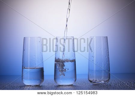 One-third full, half-filled and empty drinking glasses with reflection in drops of water on white and blue background. Water pouring into middle one.