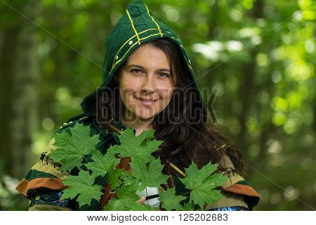 Staging photo of beautiful woman in fantasy suit with hood and leafs in forest
