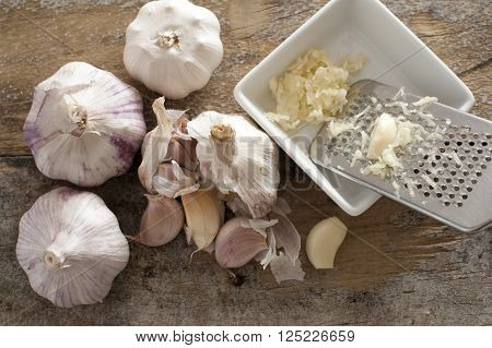 Four garlic bulbs beside a white square bowl holding shredded cloves and stainless steel grater