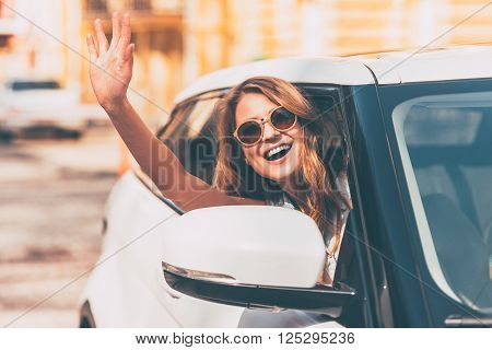 Hello! Beautiful young cheerful women looking at camera with smile and waving while sitting in her car