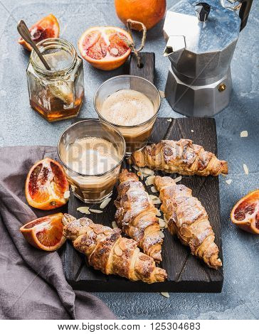 Italian style home breakfast. Latte coffee, almond croissants and red bloody oranges on dark wooden serving  board over concrete textured table, moka pot and honey jar at background, selective focus