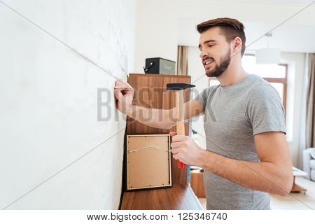 Handsome bearded young man hammering nail on the wall at home