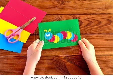 Children hands holding paper applique caterpillar. Paper sheets, scissors on a brown wooden background