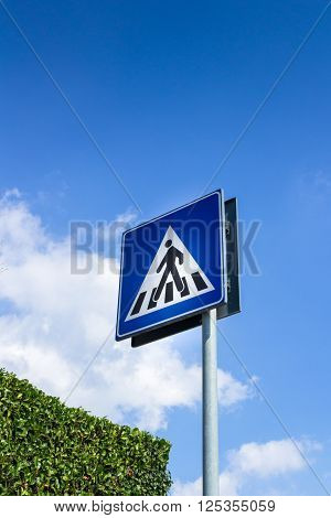 Pedestrian crossing sign with blue sky in background and green hedge.