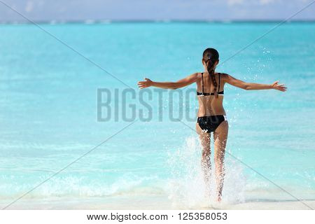 Freedom bikini woman running carefree with arms up swimming splashing water in ocean on tropical beach. Happy success girl from behind feeling free in summer Caribbean travel vacation.