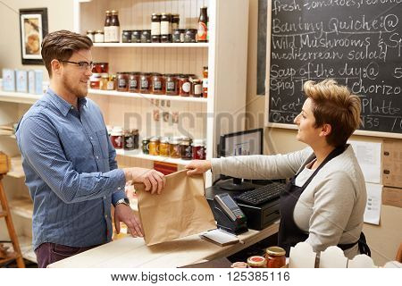 A female deli employee handing a young man a brown paper bag