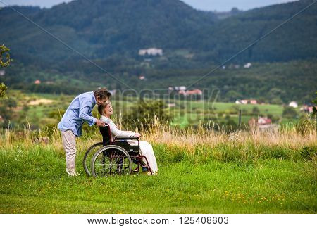 Senior man pushing woman sitting in wheelchair, kissing her, oustide in green autumn nature