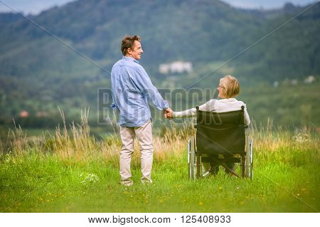 Senior man with woman sitting in wheelchair, holding hands, oustide in green autumn nature, rear view