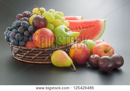 Various fresh ripe fruits arranged in a wicker basket and around on neutral gray gradient background