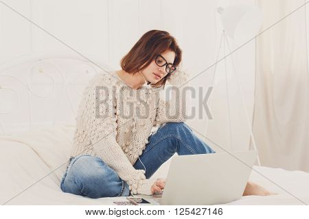 Young woman reads from laptop computer in bed. Engrossed Girl in eyeglasses and earphones with mobile phone. Attractive woman in casual clothes at white bed. Internet freelancer. High key, soft tone