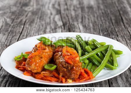 Delicious Meat Cutlets stewed with onion carrot tomato sauce decorated with basil leaves on a white dish with boiled green bean close up top view