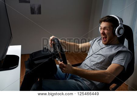 technology, gaming, entertainment and people concept - young man in headphones with pc computer playing car racing video game at home and steering wheel