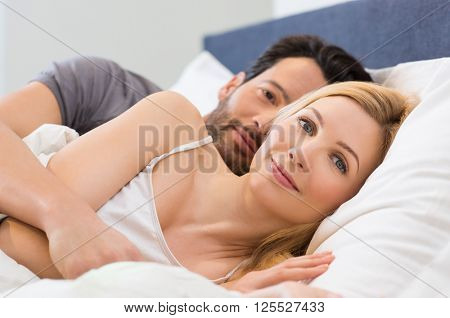 Young adult couple waking up on the bed in bedroom. Young man embracing woman while lying in bed. Happy smiling couple wake up and looking at camera. Portrait of happy woman in bed.