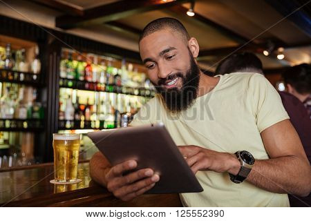 Happy african american young man drinking beer in pub and using tablet