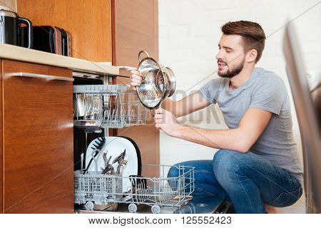 Serious bearded young man washing dishes using dishwasher on the kitchen 