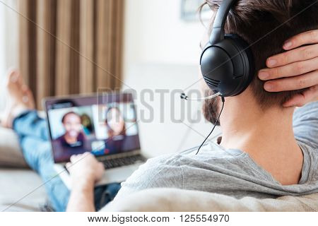 Back view of young man using headset and laptop and having videoconference at home 