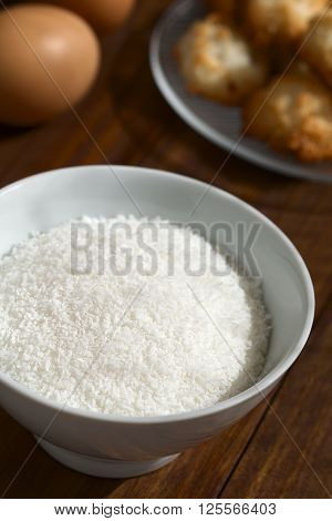 Grated coconut in bowl with coconut macaroons and eggs in the back photographed with natural light (Selective Focus Focus one third into the grated coconut)