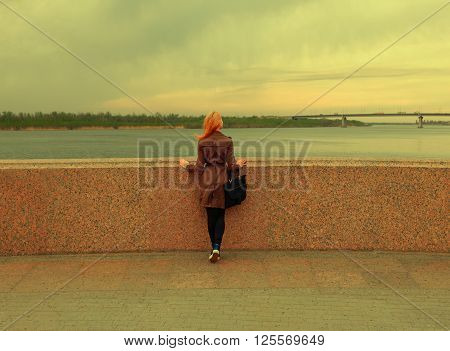 Beautiful young woman is looking at view over the river rear shot. Attractive girl enjoys summer and warm sunlight. The wind blew her hair. Vintage color, copyspace