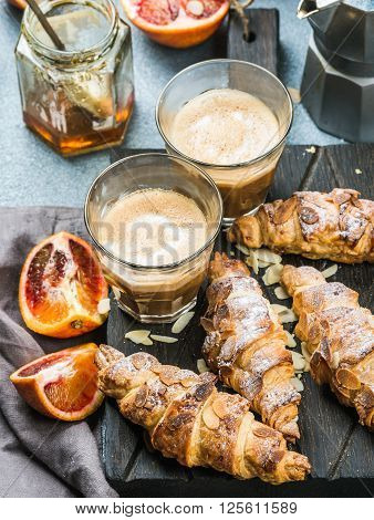 Italian style home breakfast. Latte coffee, almond croissants and red bloody oranges on dark wooden serving  board over concrete textured table, moka pot and honey jar at background, selective focus