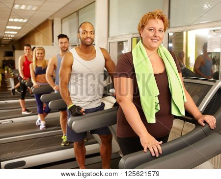 Big size woman training on running machine in gym with others.