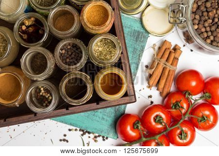 Close photo of spices and herbs small glass jars. Anise allspice cloves carry pepper chilly turmeric blue painted wooden board. Cherry tomatoes branch. Food cuisine ingredients photography.