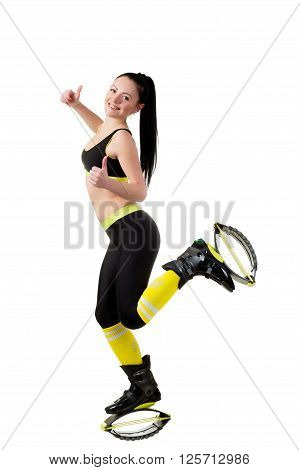 Smiling slender brunette girl with long hair in a black sportwear and kangoo jumps shoes showing a fingers up sign OK. isolated on white background in studio.