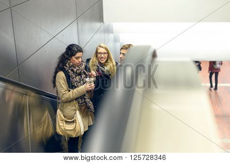 Three young adult women dressed in coat and scarves moving up on escalator in building with copy space on side