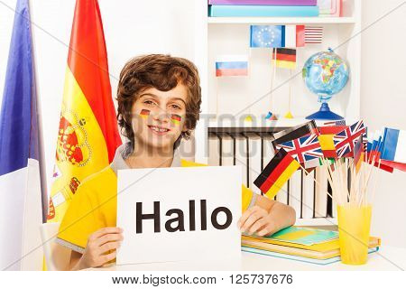 Happy schoolboy with flags on cheeks learning German, holding sign with word Hallo, at the light classroom