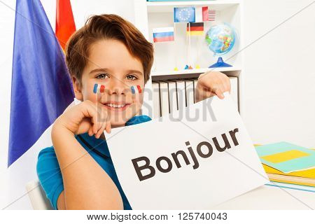 Smiling French boy holding sheet of paper with printed word bonjour - hello in French- at the classroom