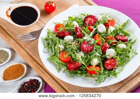 Delicious salad with tomatoes mini mozzarella arugula fried sausages on a white dish on a white rustic table caramelized balsamic vinegar in a gravy boat studio light close-up top view