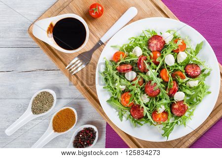 Delicious salad with tomatoes mini mozzarella arugula fried sausages on a white dish caramelized balsamic vinegar in a gravy boat on a cutting board studio light close-up top view