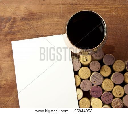 Glass of red wine shot from above on blurred wooden background with defocused wine corks from white and red wine with blank piece of white paper wine list or tasting invitation design template
