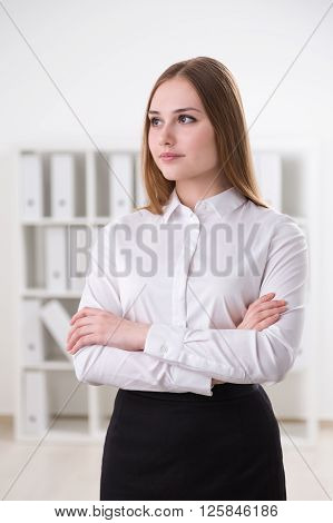 Young businesswoman standing wit hands crossed shelves at background. Office. Concept of work.
