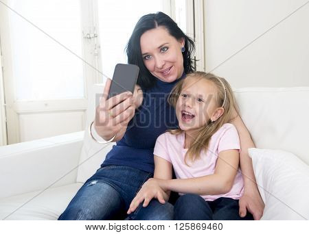 young happy woman with her little cute blond daughter taking selfie photo with mobile phone enjoying together at home sofa couch in mother and little girl self portrait picture concept
