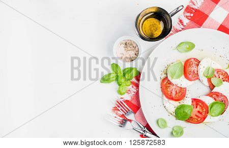 CLassic Italian Caprese salad with tomatoes, mozzarella di Buffala and fresh basil on red and white checkered napkin over white wooden background. Top view, copy space