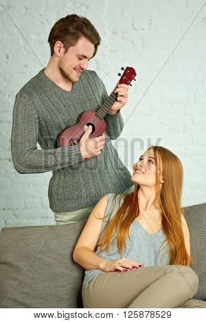 Young man playing ukulele. Beautiful woman listenin in living room.