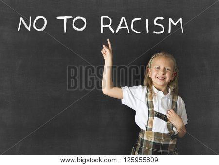 young sweet junior schoolgirl with blonde hair pointing with her finger to text no to racism written in classroom blackboard wearing school uniform in children education against racist behavior