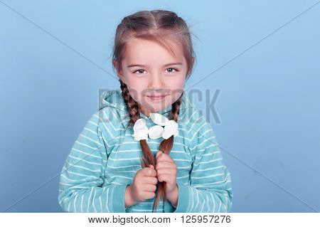 Smiling kid girl 3-4 year old posing over blue. Wearing blue striped hoodie. Holding two braids. Looking at camera.