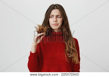 Dissatisfied young woman in casual clothes looking at her dark long hair, being displeased with work of hairdresser, frowning her face in discontent. Negative emotions and face expression