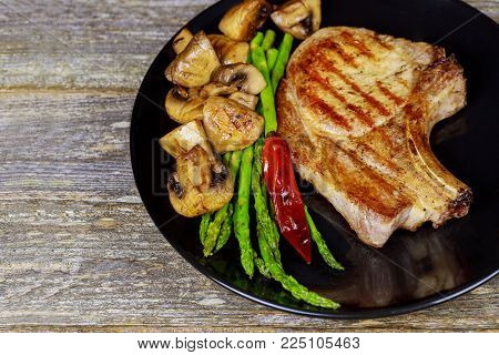 Grilled pork steaks with herbs, asparagus, garlic, mushrooms black plate background on wooden background close up.