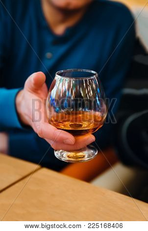 Man holding a glass of alcohol drink, man offers whiskey. A glass of whiskey in the hand of a man sitting at a wooden table