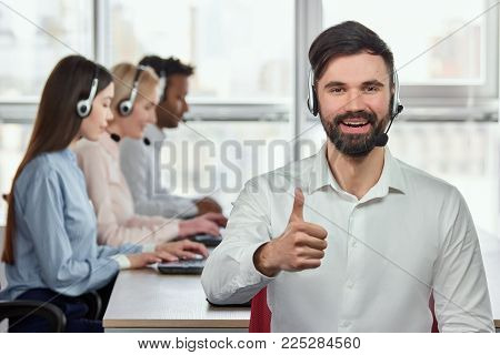 Smiling caucasian bearded man with thumb up in bright office. Cheerful young handsome male call center opearator shows thumb up against blurred co-workers background in bright office.