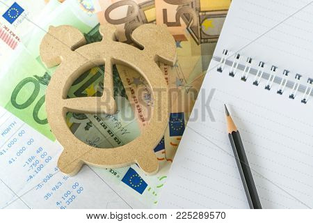 Time for invest, tax and debt calculation, pencil on white notepad paper with wooden alarm clock on Euro money and salary slip.