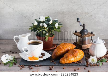 Morning set with two croissants, cup of aroma coffee, jug of cream to coffee and azalia on a gray wooden background.