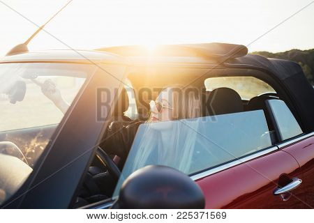Pretty and good looking young blonde woman or model, sits inside convertible cabriolet car, before opening roof, smiles during beautiful sunset. proud new car owner teenager dream come true