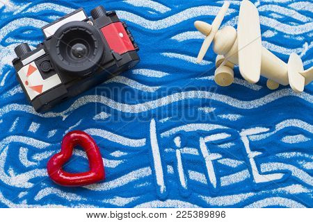 Life Inscription On A Blue Colored Sand With Waves, Roll Film Camera And Wooden Plane, Top View, Fla