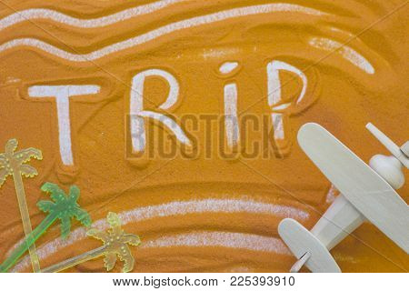 Trip Inscription On An Orange Colored Sand With Waves, Plastic Palm Trees And Toy Wooden Airplane, T