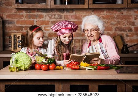 Granny with two granddaughters reading recipe from cooking book and cooking salad