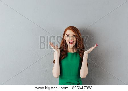 Photob of excited happy redhead young lady in green dress standing over grey wall background. Looking camera.