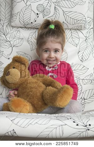 Portrait Of Emotional Little Girl Is Sitting On A Chair And Hugging A Bear Toy